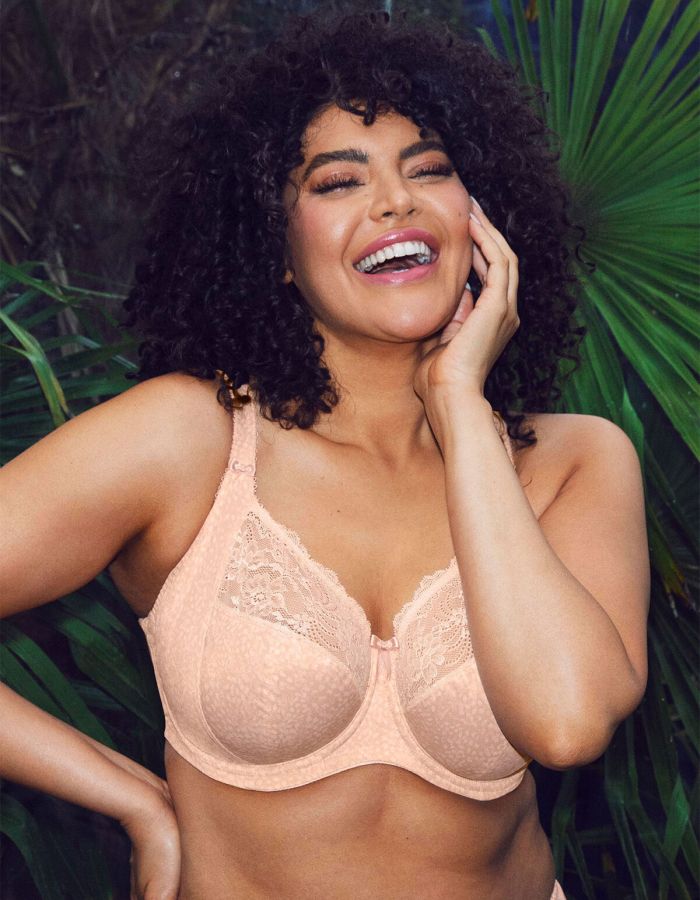 Woman wearing a peach underwired lace bra, smiling against a leafy green background.