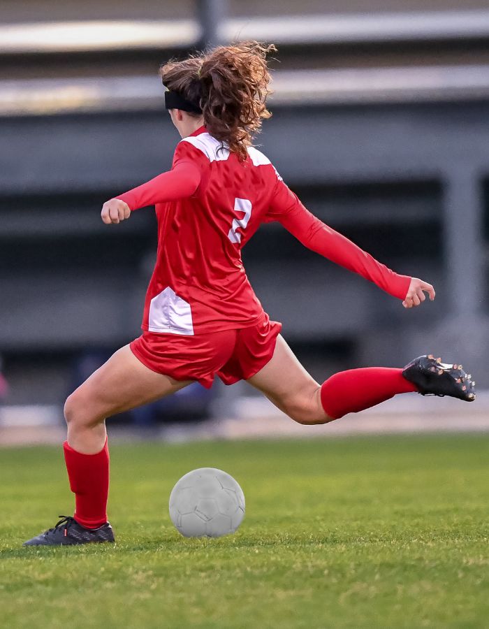 Female soccer player in red uniform running and kicking a ball on a grass field during a competitive football match.