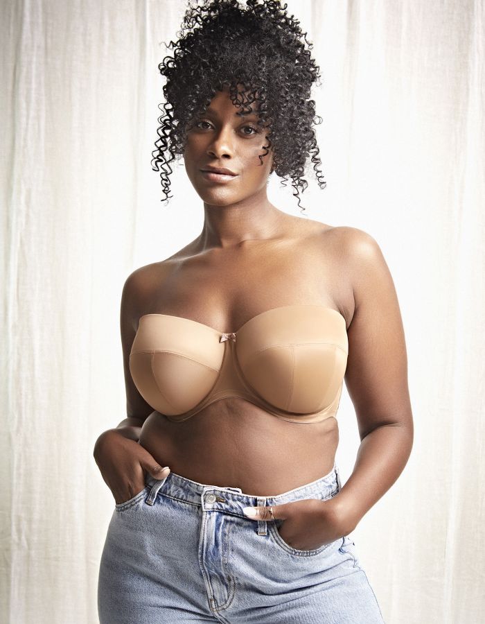 Woman wearing a nude strapless bra with supportive full cups and light blue jeans against a softly lit neutral background.