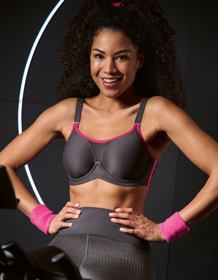 Woman wearing a grey underwired sports bra with pink trim, supportive cups and wide straps in a gym setting.